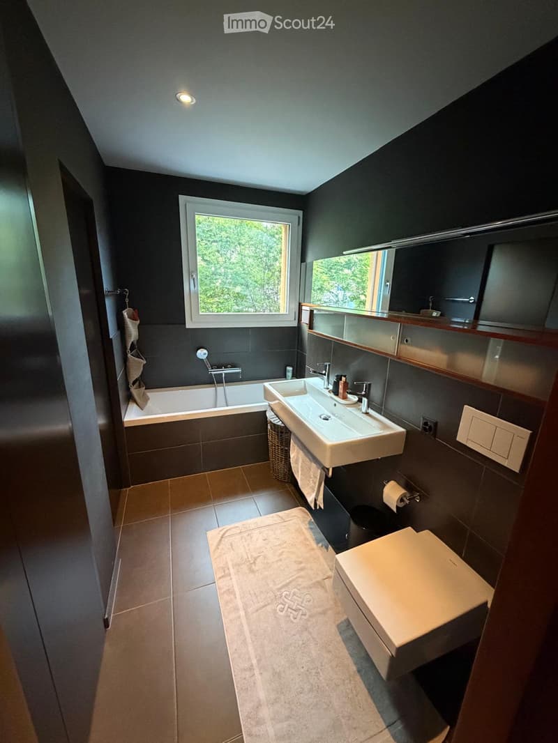 Modern bathroom with dark grey walls, a bathtub, a double sink, a toilet, a large mirror, and a towel on the floor.