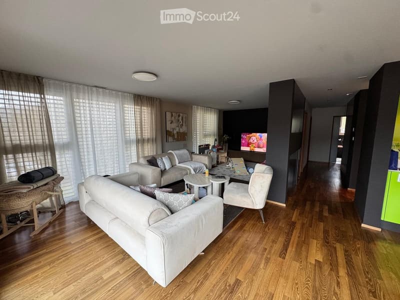 Modern living room with hardwood flooring, large windows with sheer curtains, and contemporary furniture including a sofa, chairs, coffee table, and a TV screen.