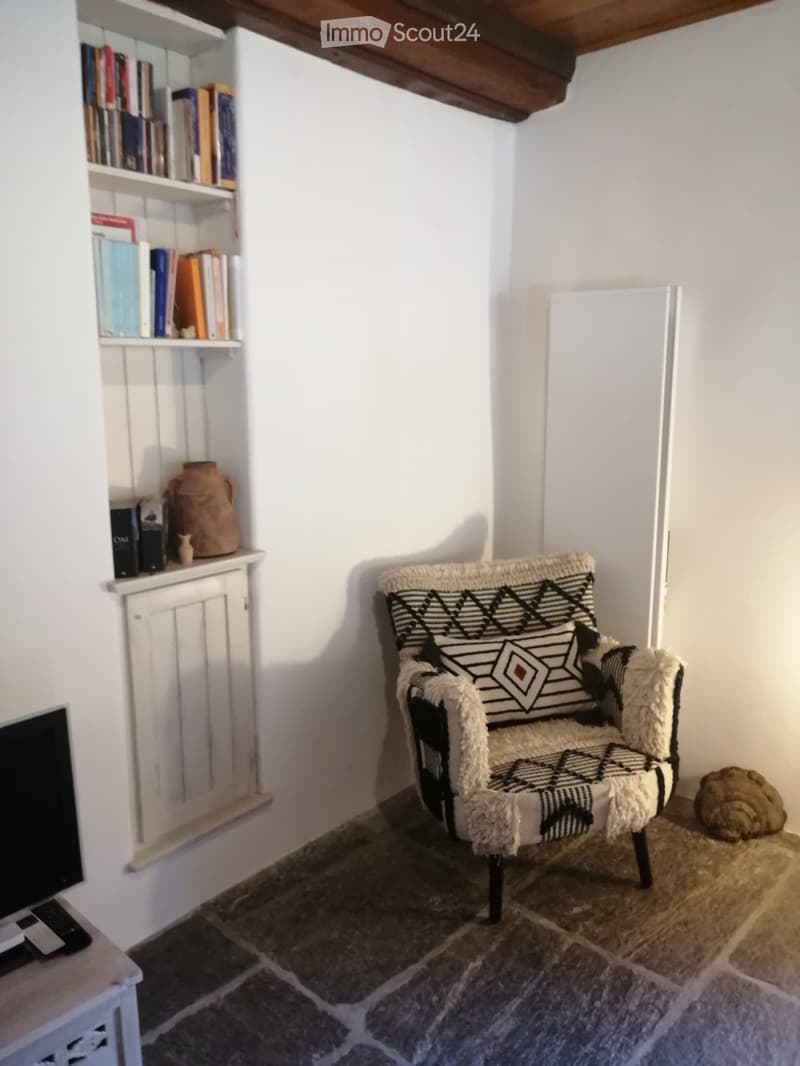 A cozy, small room with a white wall, a wooden ceiling beam, a patterned armchair, a shelf with books and a decorative vase, and a stone-tiled floor.