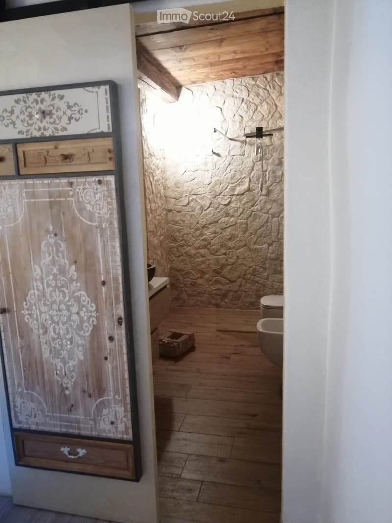 Bathroom with stone walls, wooden floor, toilet, bathtub, and a wooden cabinet with decorative carvings.