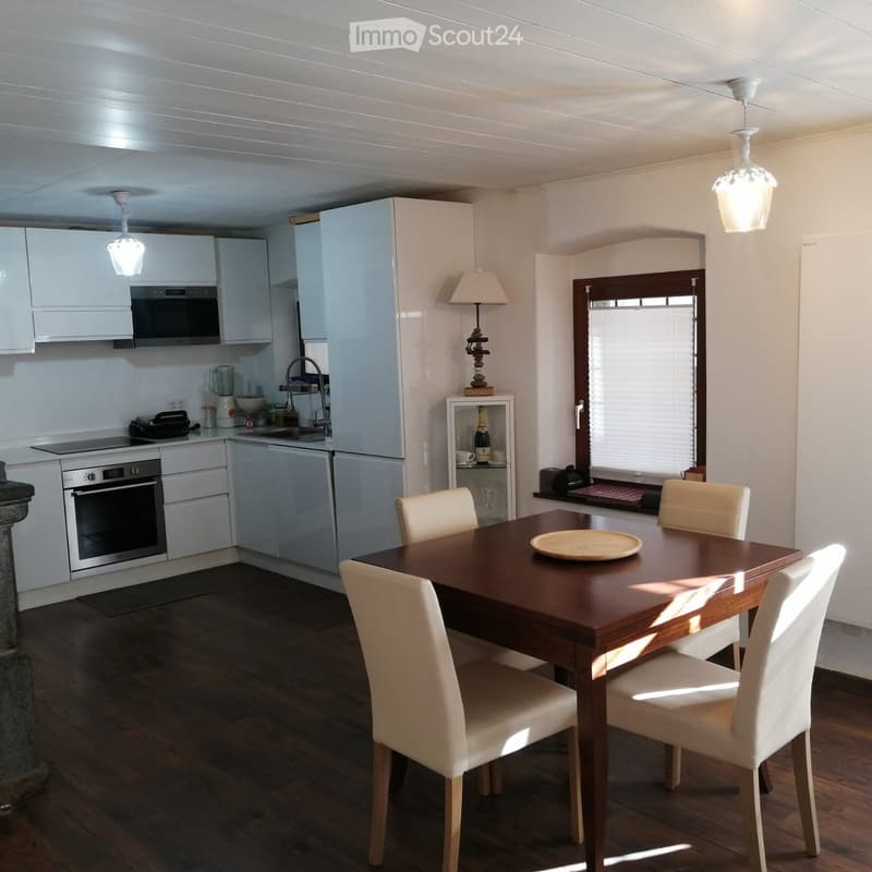 Modern kitchen with white cabinets, stainless steel appliances, a wooden dining table with four chairs, and a small shelf with decorative items.