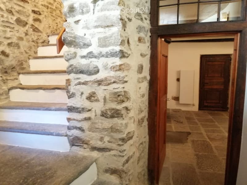 A narrow stone-walled corridor with wooden doors and a staircase leading upwards, featuring a tiled floor and a small window at the top.