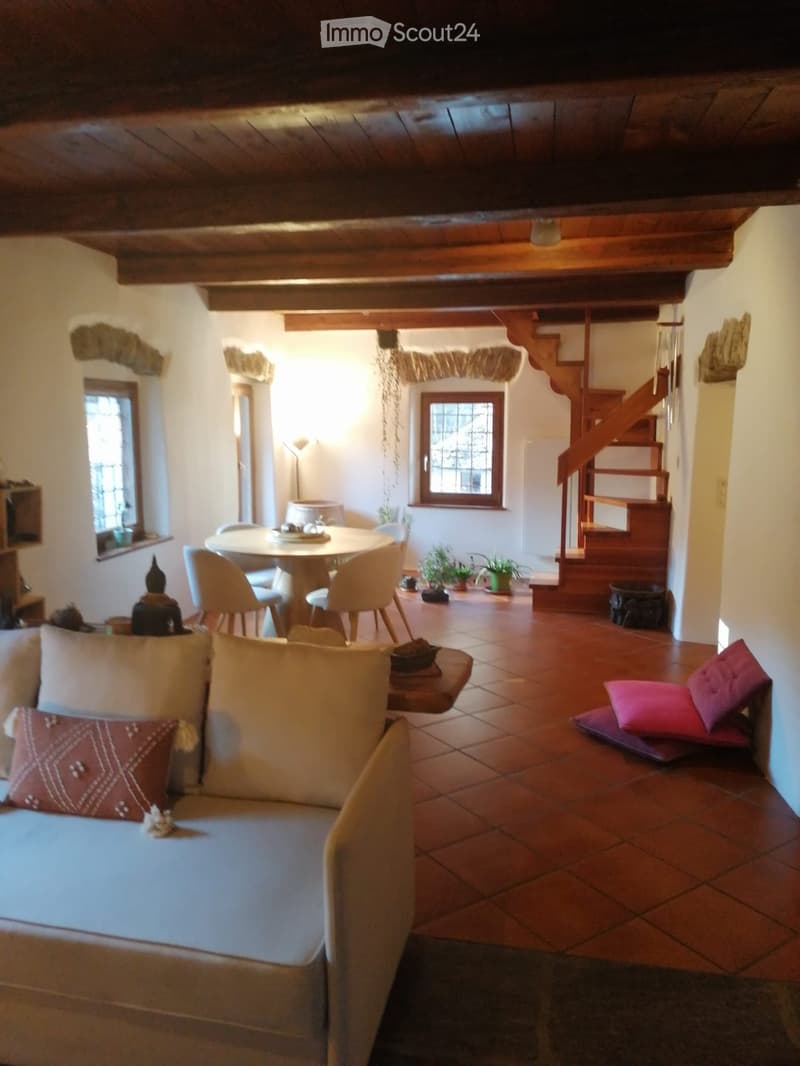 Living room with exposed wooden ceiling beams, terracotta tiled floor, sofa, small table, chairs, and a staircase leading to an upper level.