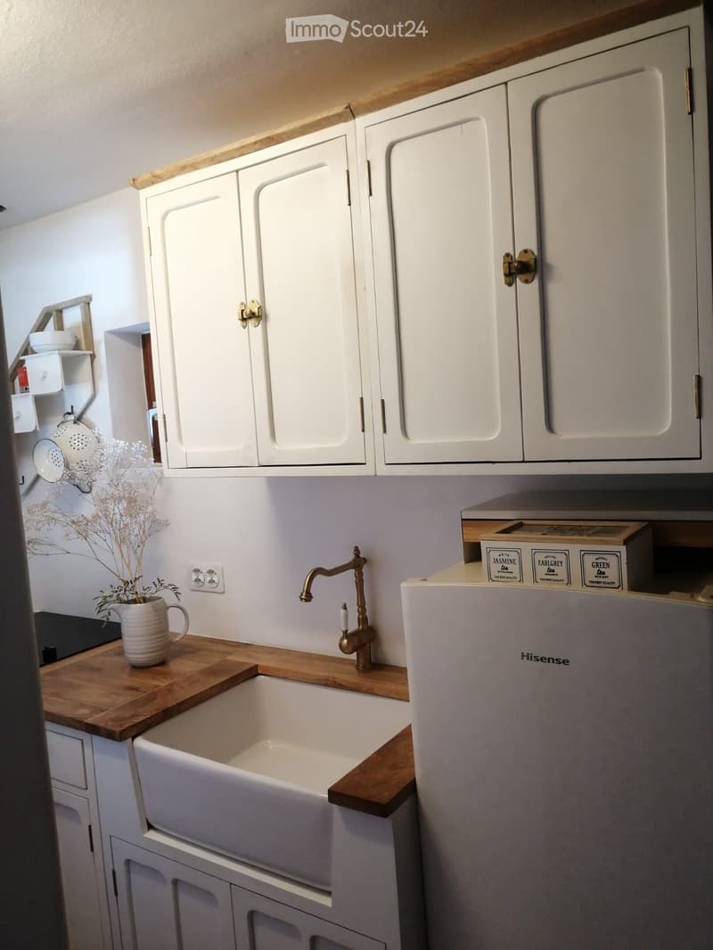 A modern kitchen with white cabinets, a wooden countertop, a sink with a brass faucet, and a Hisense refrigerator. A vase with dried flowers is placed on the countertop.