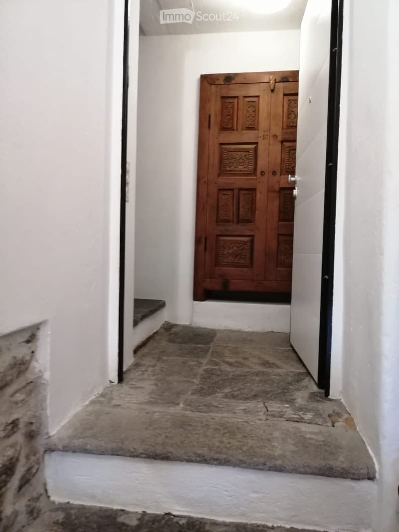 A narrow, white-walled corridor with a wooden double door at the end and a stone floor.