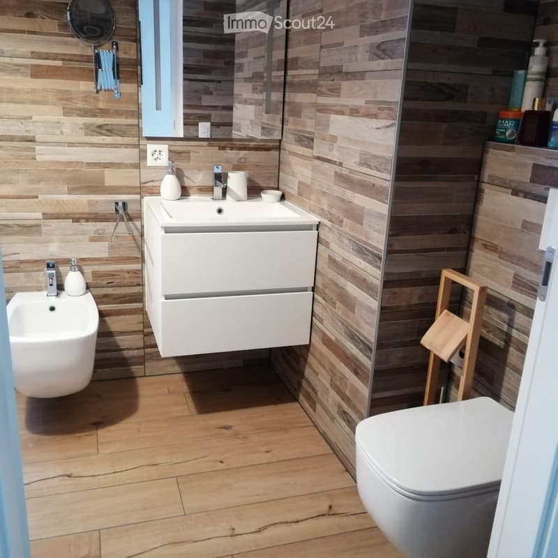 Modern bathroom with wooden walls and floor, white sink, toilet, mirror, and small wooden stool.