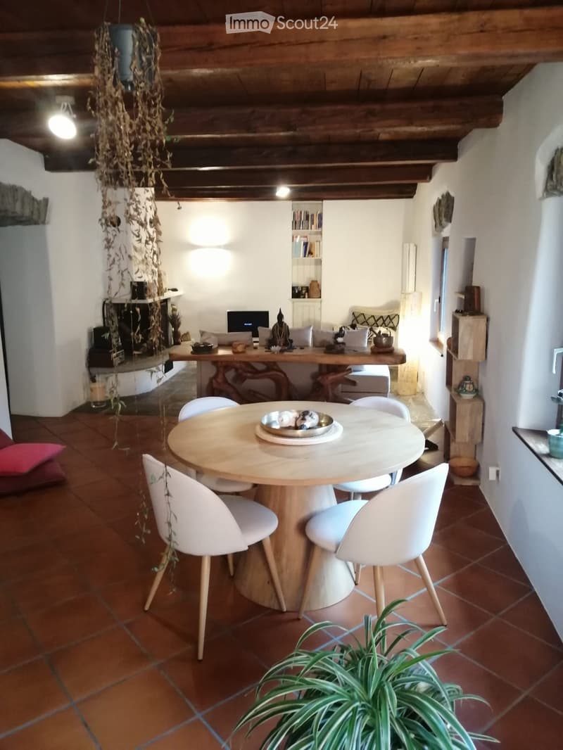 A cozy living and dining area with exposed wooden beams, a round dining table with four white chairs, a sofa, a fireplace, and a bookshelf. The room features terracotta tiles and a hanging plant.