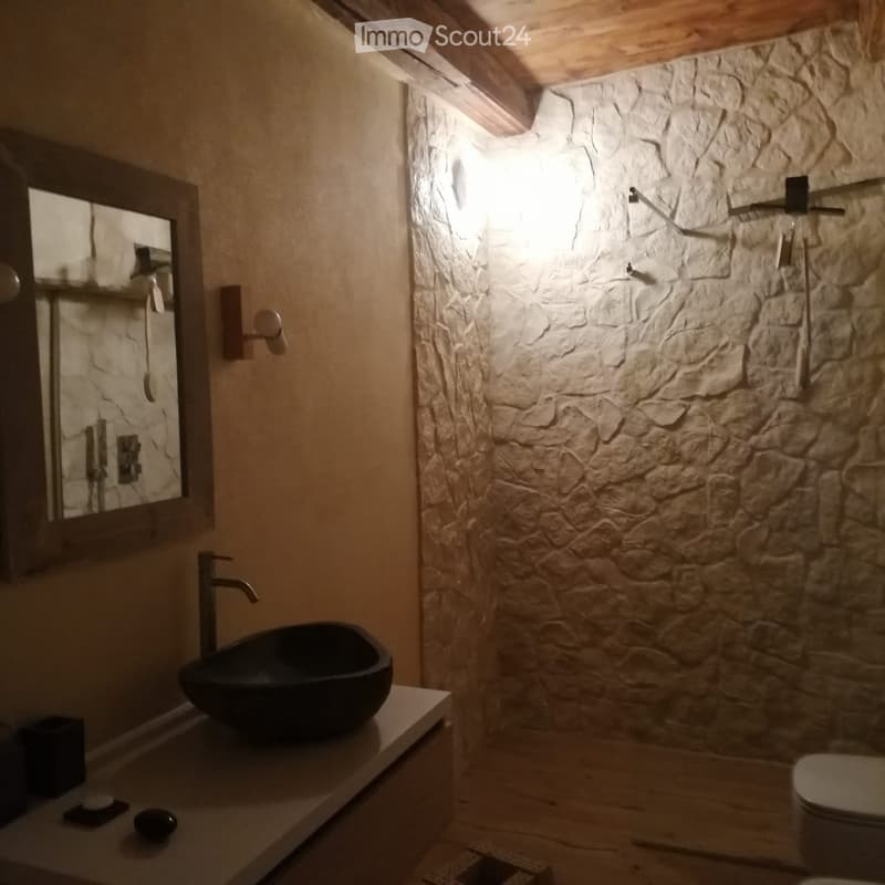 Modern bathroom with a stone wall, black sink, and a sleek faucet. Features a large mirror and a toilet.