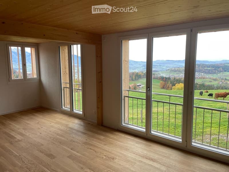 Bright, empty room with wooden flooring and large windows offering a scenic view of a green valley and mountains.