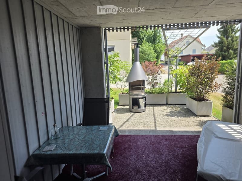 Balcony with a table, chair, and bottle, overlooking a garden with a pizza oven and potted plants.