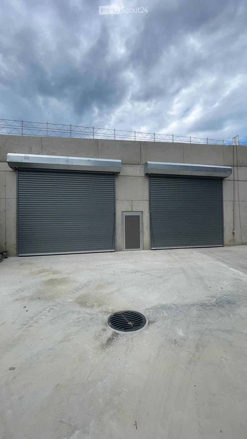 Concrete building with two large roll-up garage doors and a small access door, topped with a metal awning and a security fence on the roof.