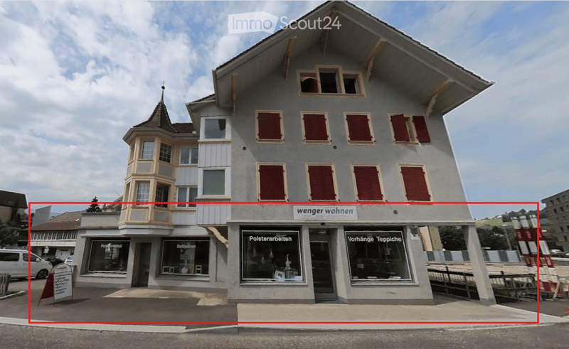 3 story building, gray exterior, red shutters, storefronts on the ground floor, sign 'wenger wohnen', sign 'Polsterarbeiten', sign 'Vorhänge Teppiche'