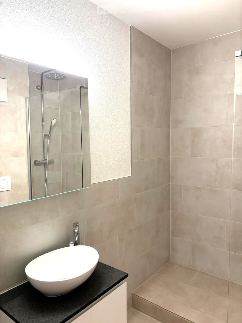 Modern bathroom with a white sink, black countertop, glass-enclosed shower, and beige tiles.