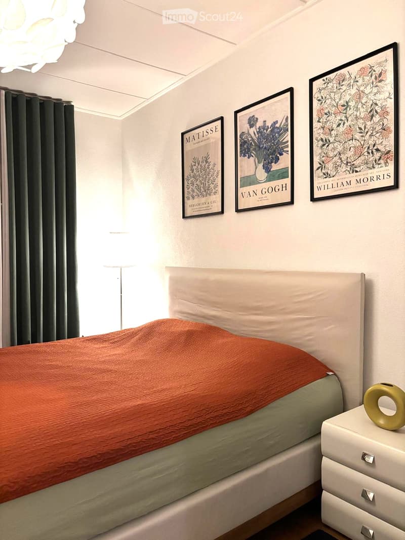A modern bedroom with a neatly made bed, framed artwork, a nightstand with a decorative vase, and a floor lamp near the window.