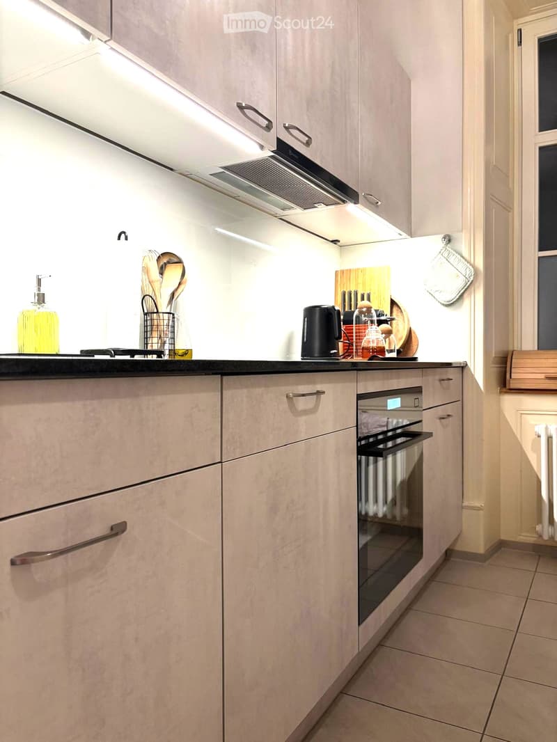 Modern kitchen with light-colored cabinets, stainless steel handles, and a built-in oven. Countertop features a bottle of olive oil, dish soap, and a basket of kitchen utensils. A wooden cutting board with knives and a black kettle are also visible.
