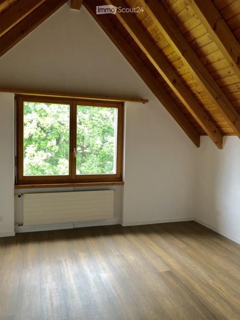 Empty attic room with wooden slanted ceilings, wooden flooring, and a large window with wooden frame showing greenery outside. A white radiator is placed below the window.