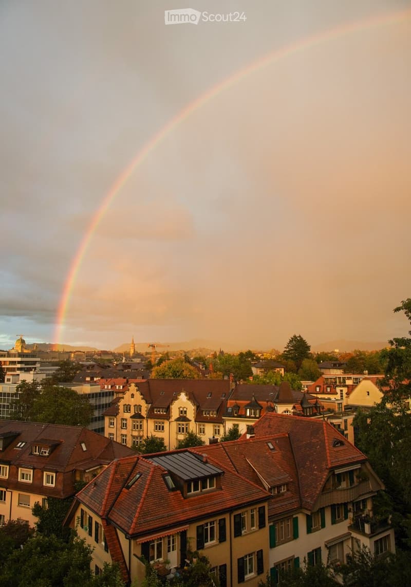 A panoramic view of a cityscape with multiple residential buildings featuring red-tiled roofs and classic European architecture, surrounded by trees and a visible rainbow in the sky.