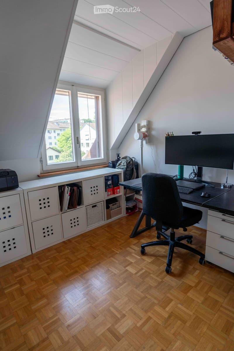 Bright attic office with wooden herringbone flooring, large windows, modern desk setup, ergonomic chair, and storage unit.