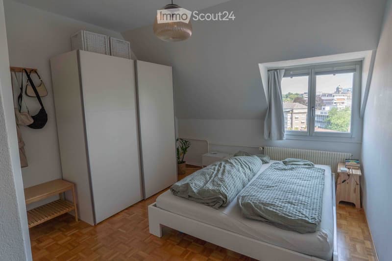 Cozy bedroom with a double bed, nightstand, potted plant, large wardrobe, and a bench near the door. The room features wooden flooring and large windows with curtains, offering a view of the city.