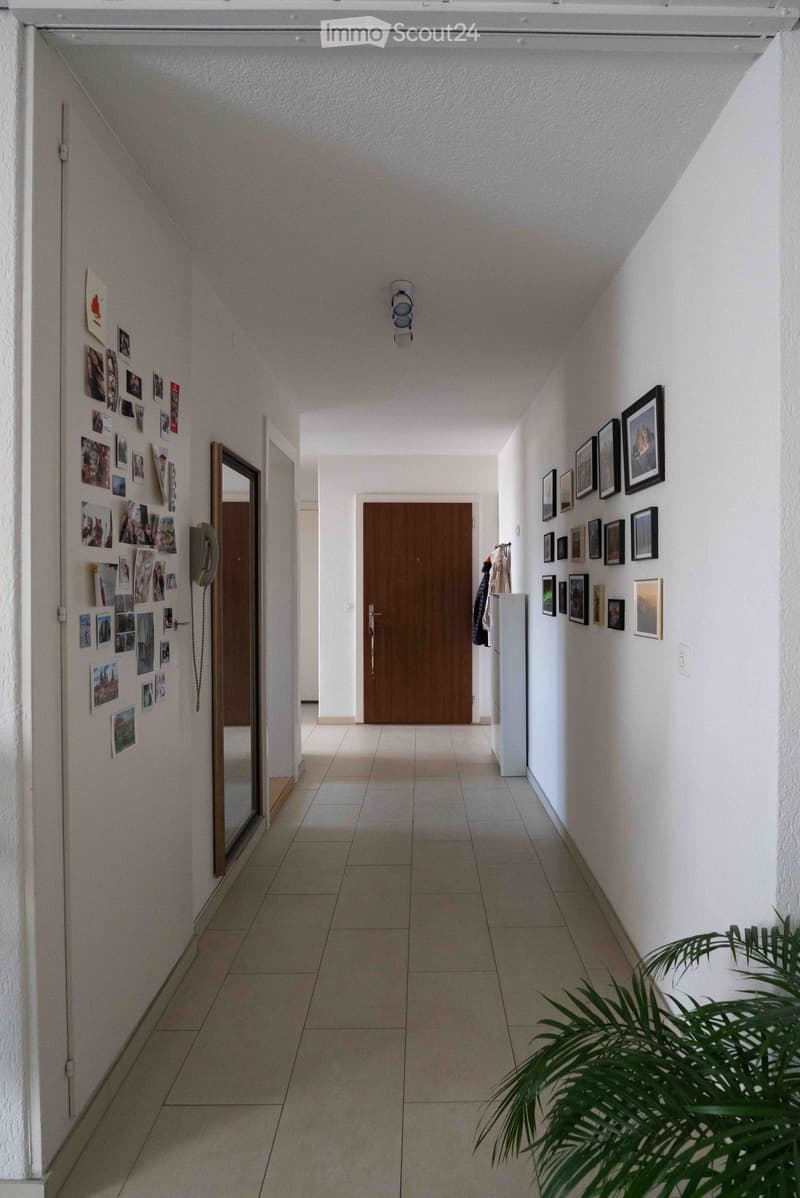 A long, narrow hallway with tiled flooring, white walls, and framed pictures on both sides. A wooden door is visible at the end, and a mirror is mounted on the left wall.