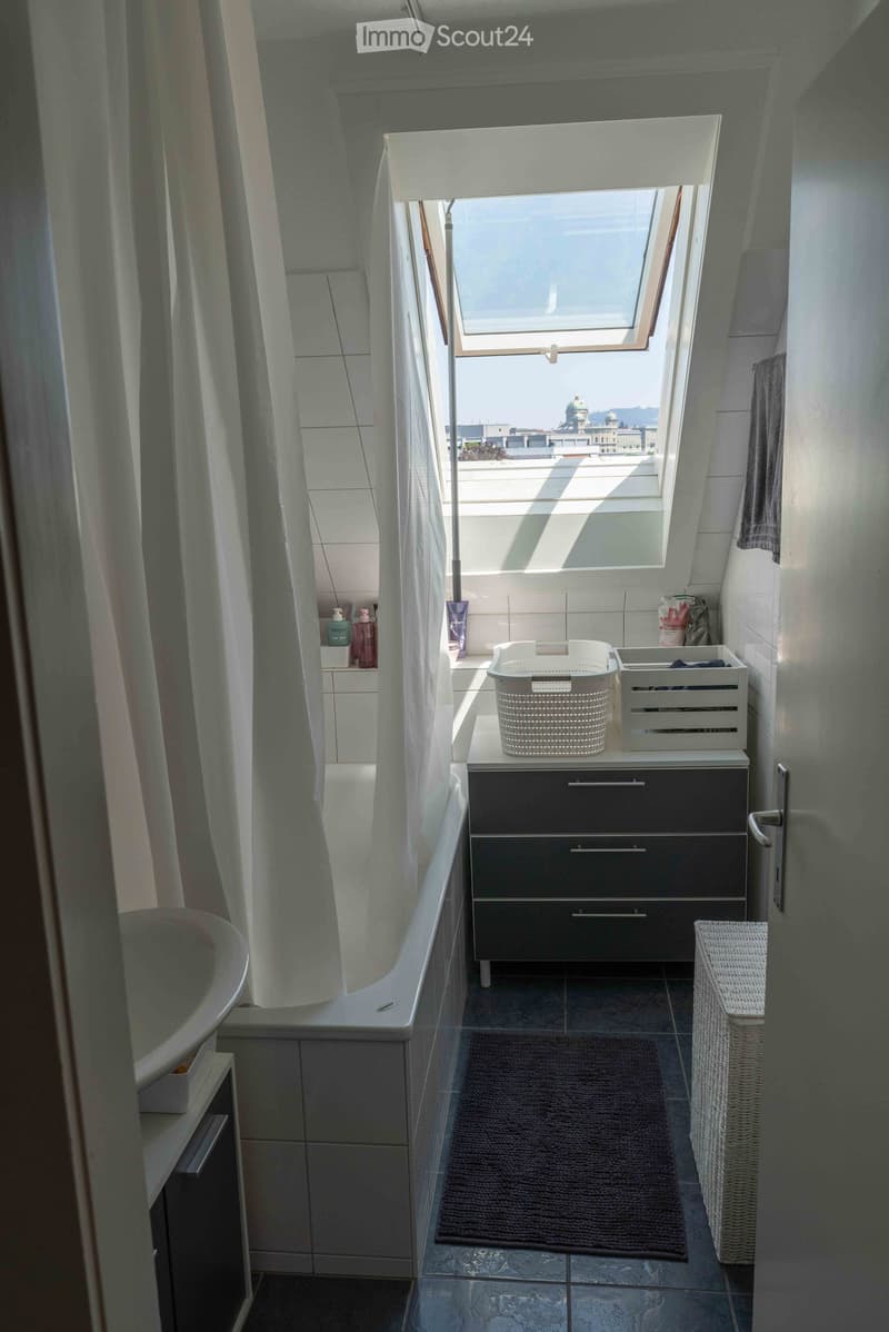 Modern bathroom with a bathtub, sink, and a large skylight window offering a city view. Includes a dark-colored cabinet, a white basket, and a towel on the floor.