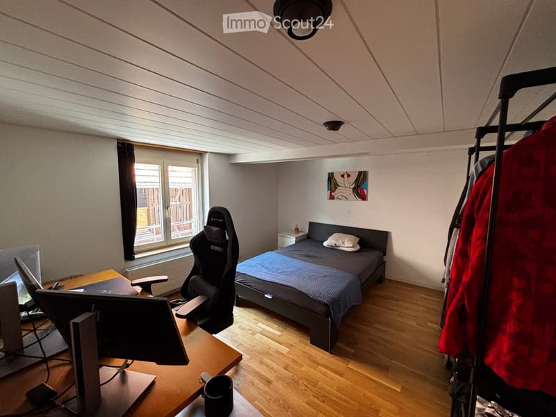 Bedroom with a single bed, desk with laptop and screen, gaming chair, and a coat rack with jackets.