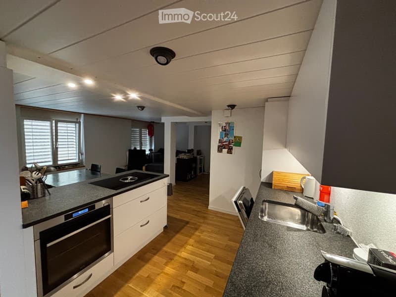 Modern kitchen with white cabinets, grey countertops, built-in oven, microwave, sink, and various appliances. The space features wooden flooring and ample natural light from large windows.