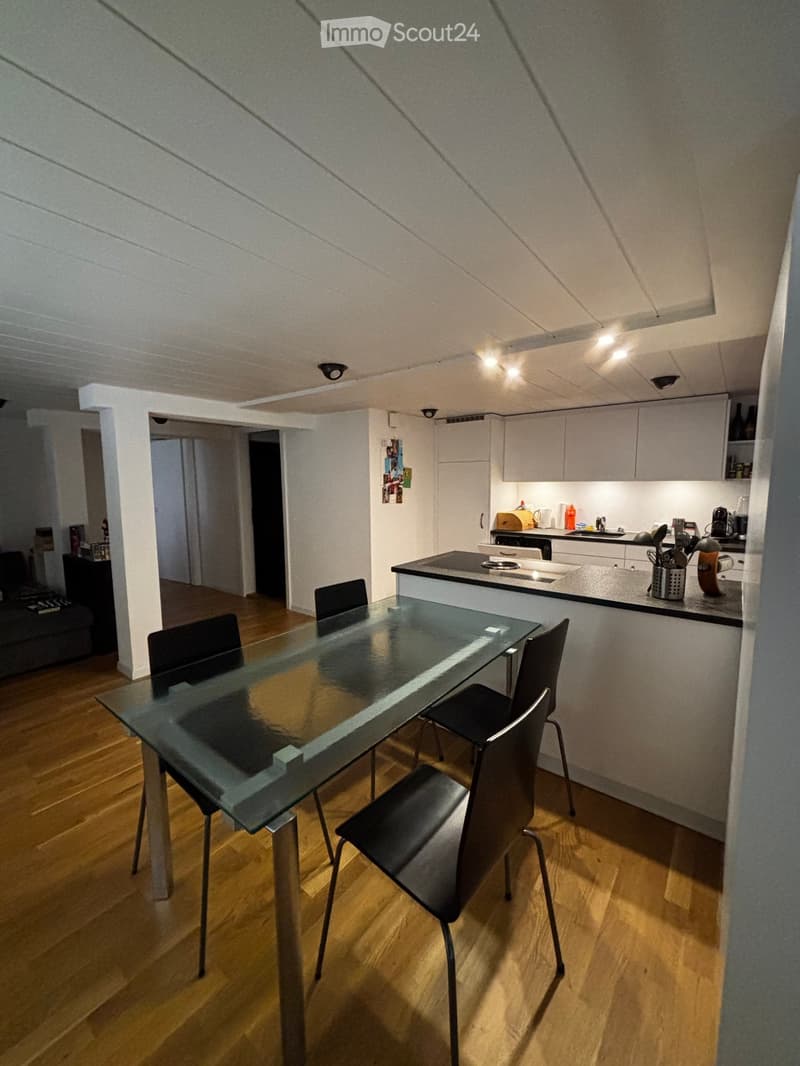 Modern, open-plan living and dining area with a glass dining table and chairs, a kitchen area with cabinets, a counter, and various appliances.