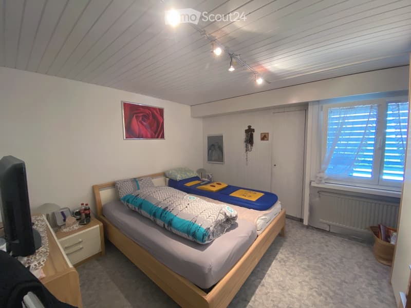 Cozy bedroom with a lofted bed, wooden flooring, white walls, and a window with blinds. Features include a picture frame of a rose on the wall and a small cabinet.