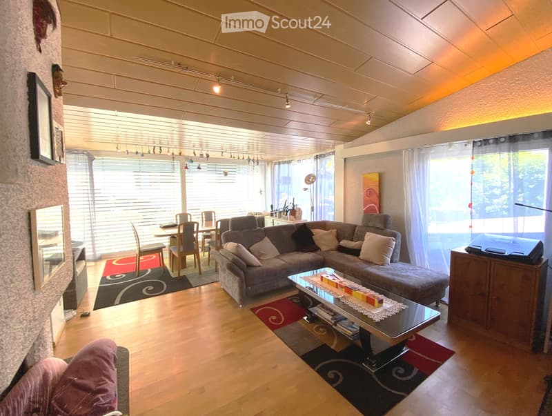 A spacious living room with a raised platform sofa, a dining table with chairs, large windows, and a wooden floor.