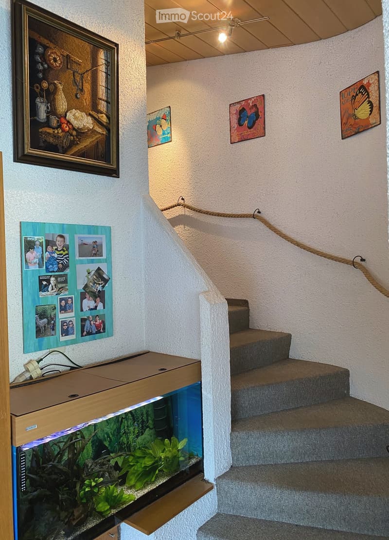 A narrow hallway with a staircase leading upwards, featuring a fish tank on the left, framed artwork on the walls, and decorative tiles on the floor.