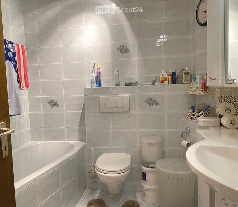 A modern bathroom with white tiles, a sink, a mirror, a toilet, and various toiletries including toothbrushes and toothpaste. A towel hangs on the side, and a small cabinet is visible.