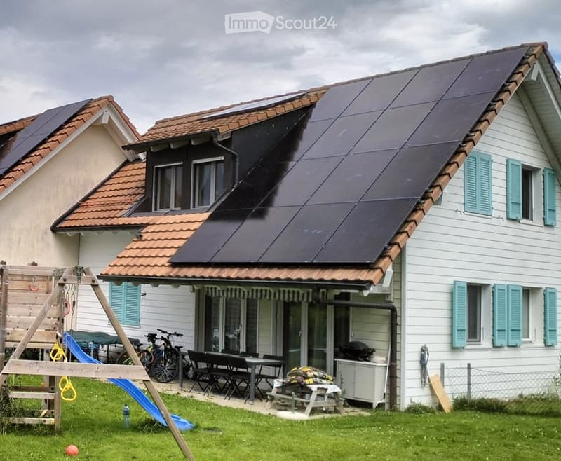 Two-story house with white siding, teal shutters, and solar panels on the roof. The front yard includes a playground with a slide, swings, and a small table with chairs.