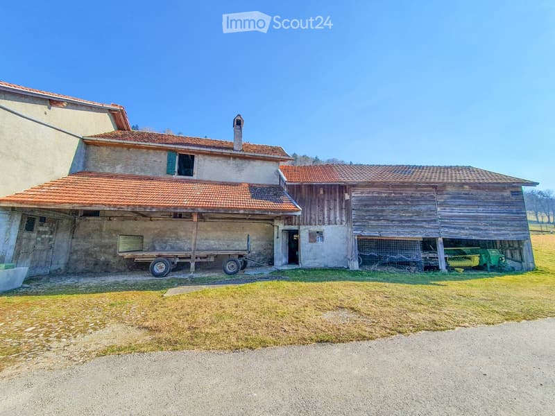Two-story rustic building with red-tiled roof, stone and wooden walls, and a covered wooden outbuilding with agricultural equipment.