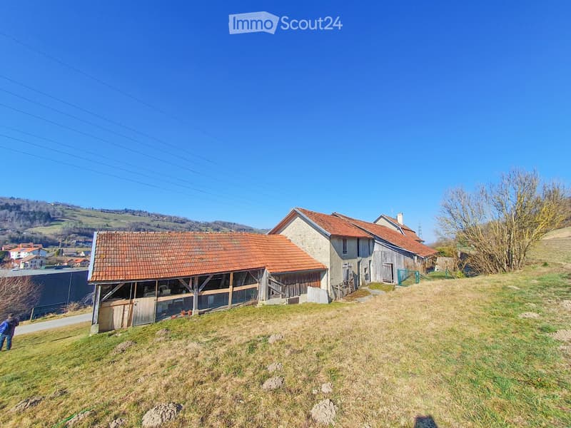 A two-story house with a red-tiled roof, stone walls, and a wooden outbuilding, situated on a grassy slope with a rural backdrop.