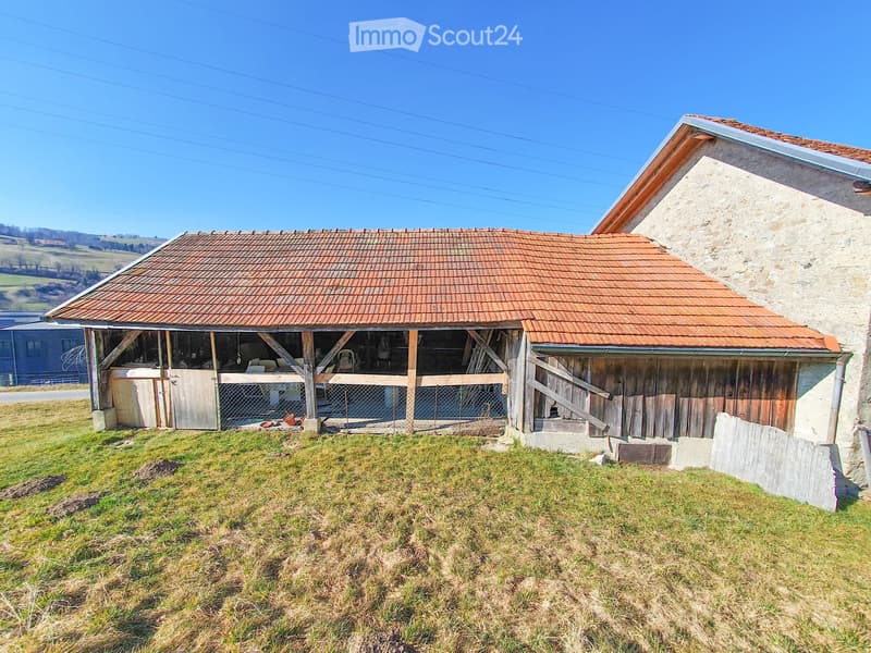 A rustic, two-story building with a red-tiled roof and stone walls, featuring a wooden stable or shed attached to the main structure. The surrounding area is grassy with a clear blue sky in the background.