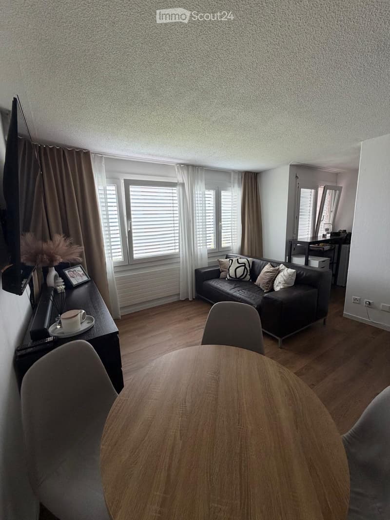 Modern living room with wooden flooring, large windows, a black leather sofa, a round wooden table, and a side table with a cup and decorative items.