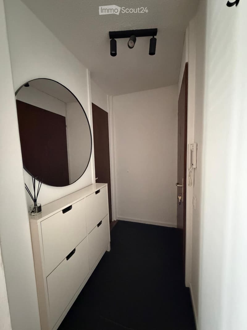 A narrow hallway with white walls, black carpet, a white cabinet with drawers, a round mirror, and a small decorative item on top of the cabinet. The hallway has a modern design with recessed lighting and a door at the end.