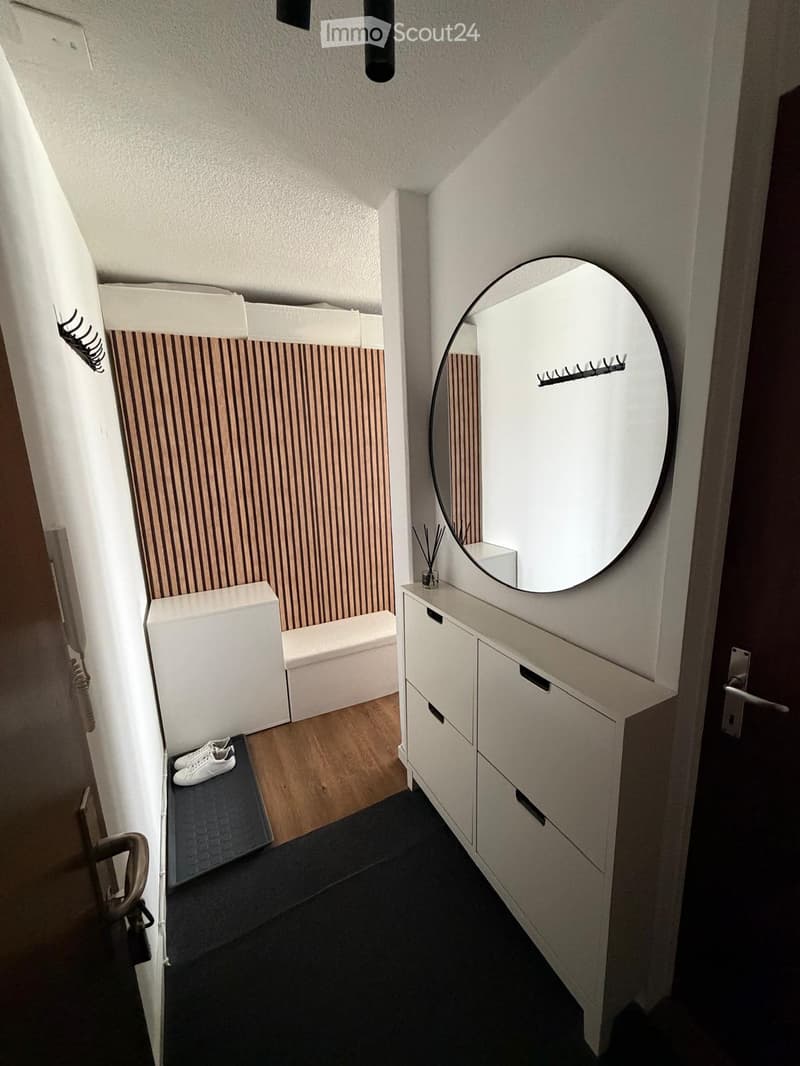 Modern hallway with a sleek vanity, round mirror, and a small shoe rack. The space features a wooden floor and white walls with subtle striped accents.