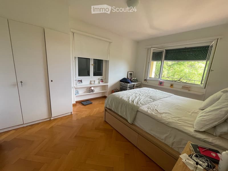 A well-lit bedroom with wooden flooring, a large bed with white bedding, a nightstand with a lamp, and a shelf with picture frames and decorative items.