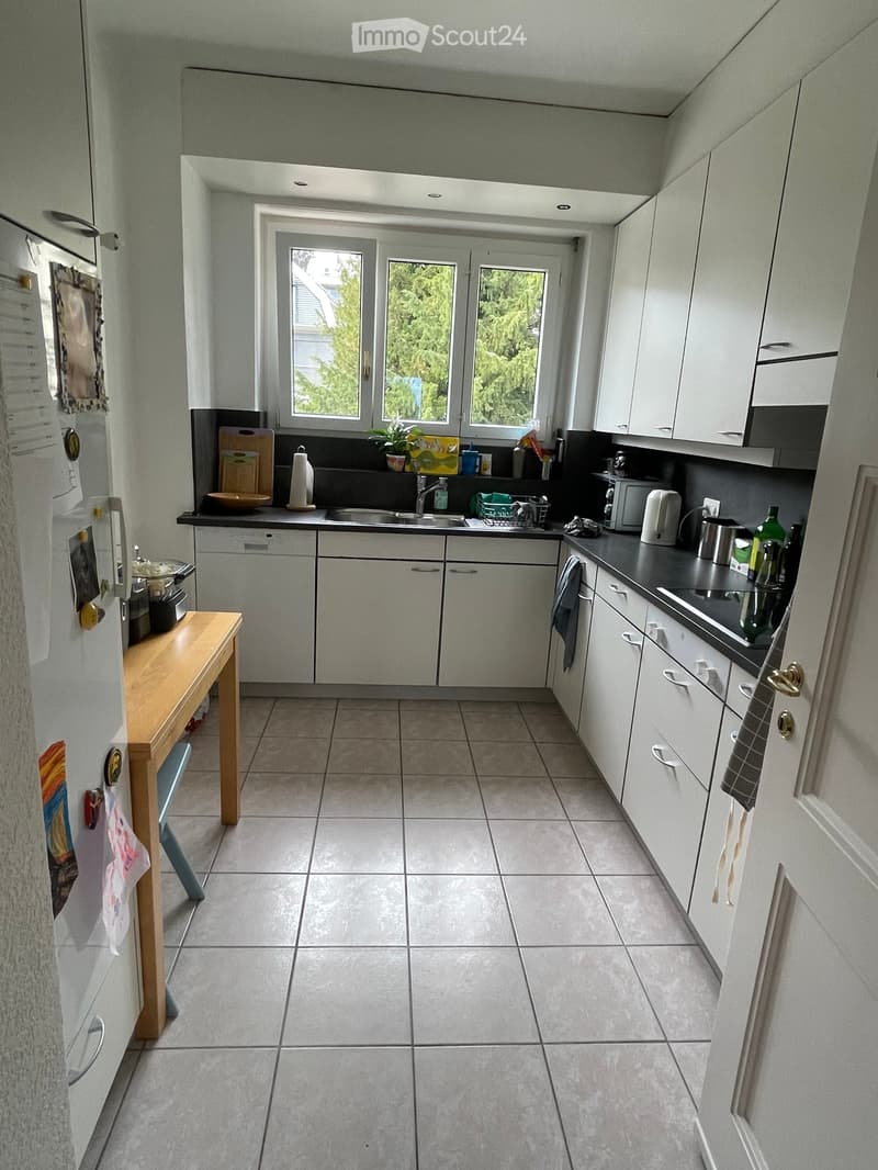 Modern kitchen with white cabinets, tiled floor, and various appliances including a refrigerator, microwave, and oven. There is a wooden table near the refrigerator and a sink with a faucet in the center.