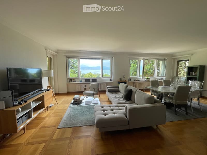 Bright living room with large windows offering a scenic view, featuring a grey sectional sofa, coffee table, dining table with chairs, TV, and floor lamp.