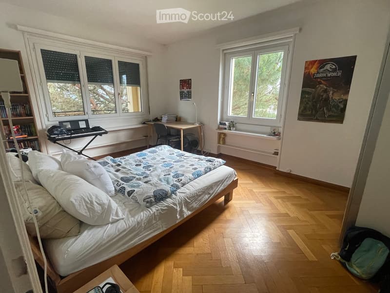 Cozy bedroom with wooden flooring, large windows, a neatly made bed, a desk with a chair, a fan, and a bookshelf.
