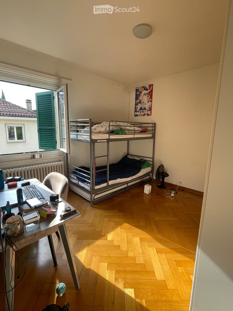 A small bedroom with wooden flooring, a bunk bed, a desk with a laptop, and a fan near the window.