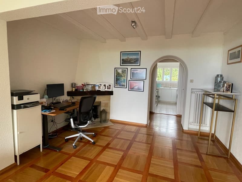 A well-lit study or office space with wooden flooring, a desk setup including a monitor, keyboard, mouse, and printer, a comfortable office chair, framed pictures on the wall, and a small table with decorative items. An arched doorway leads to another roo