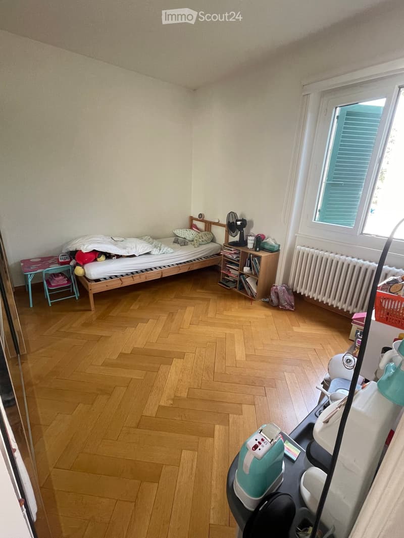 A small bedroom with wooden flooring, a single bed with pillows and blankets, a nightstand with a fan, a small table with a chair, a radiator under a window, and a shelf with books and other items.