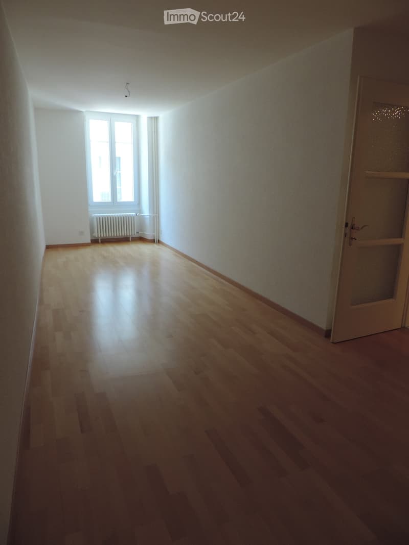 Empty room with wooden flooring, white walls, and a window with a radiator below it. A door is visible on the right side.