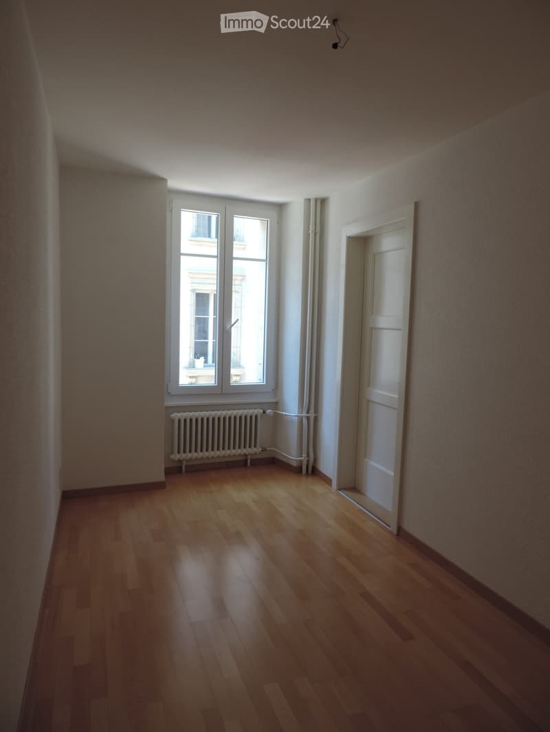 Empty room with wooden flooring, white walls, a window with double glass panes, a radiator, and a closed white door leading to another space.