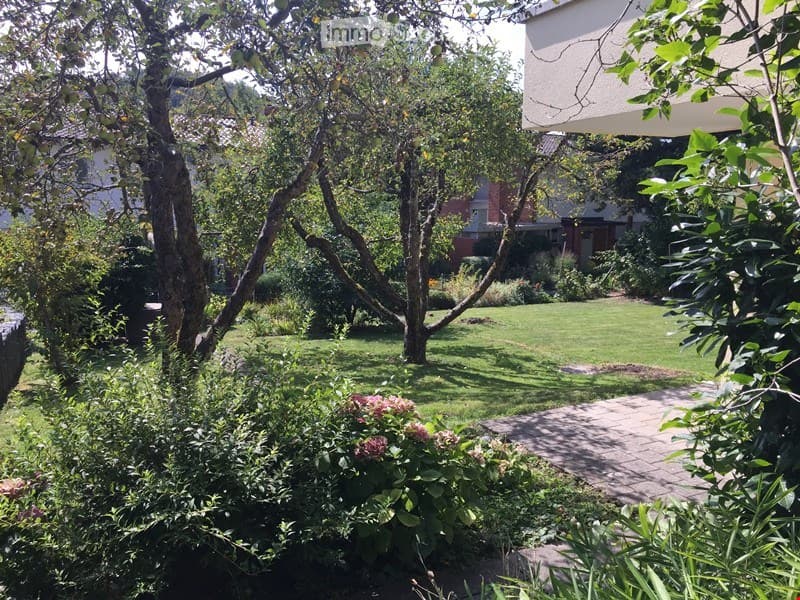 Well-maintained garden with a central lawn, mature trees, and a paved pathway. Surrounding area includes shrubs and flowering plants.