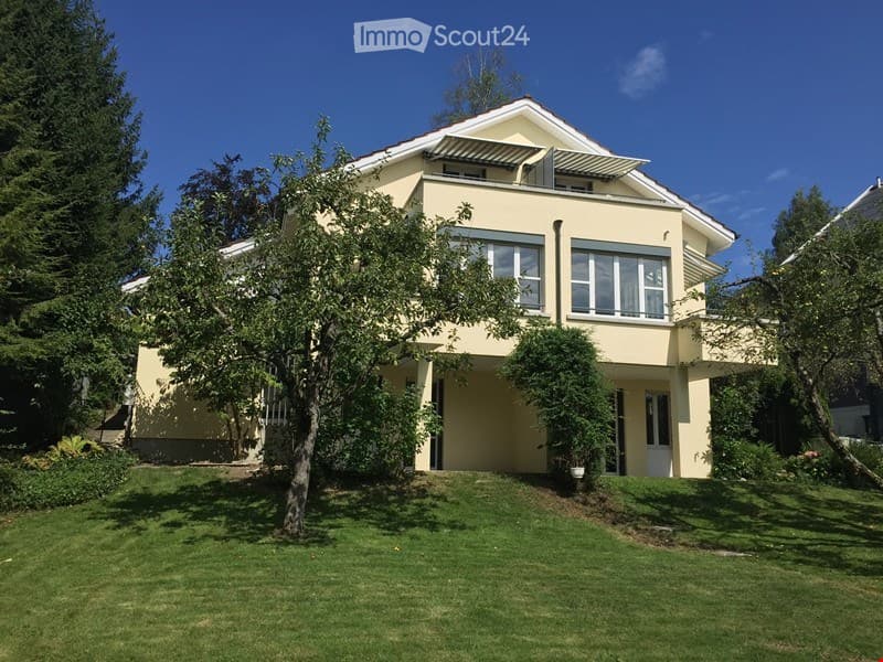 Two-story house with a light yellow exterior, large windows, and a balcony on the upper floor. The house is surrounded by a well-maintained lawn and trees.